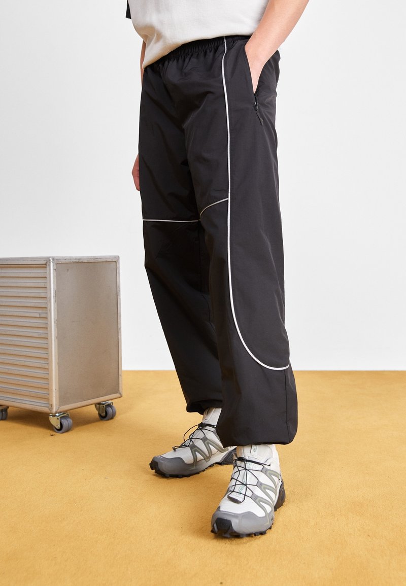 Mustat nylonhousut, joissa on joustava vyötärönauha, valkoiset reunalistat, sivussa vetoketjutasku ja harmaat vaelluskengät keltaisella lattialla.