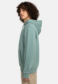 Sudadera verde menta hecha de tela suave, con un bolsillo frontal, puños acanalados y una capucha con cordón. Usada sobre pantalones de color claro.