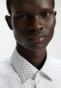 Camicia a maniche corte con tessuto bianco e un sottile motivo geometrico blu. Include un colletto a punta e chiusura con bottoni.