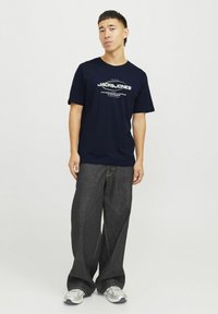 Mørkeblå bomulds t-shirt med hvidt trykt logo. Løs pasform i mørkegrå denim bukser og grå sneakers med hvide detaljer.