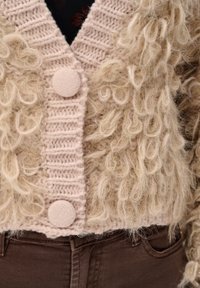 Beżowy gruby sweter kardigan z teksturą pętelkowej przędzy i guzikami pokrytymi tkaniną, noszony na brązowych dżinsach.