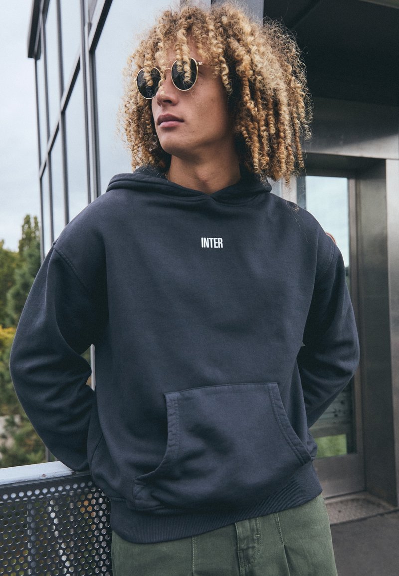Sual de marinha com "INTER" impresso em branco, apresentando um bolso frontal, capuz com cordão e punhos canelados. O modelo usa óculos de sol e calças de cor oliva.