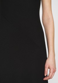 Gros plan sur le bras et la main d'une personne à côté d'un vêtement en tissu noir côtelé avec des détails de coutures diagonales.