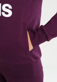 Sudadera morada con texto blanco, con un bolsillo frontal y puños acanalados. Hecha de una tela suave con una textura lisa.