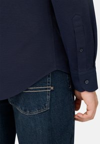 Temno modra teksturirana srajca z gumbi na manšetah, v kombinaciji s temno modrimi jeansi z svetlimi šivi in vidno blagovno znamko na žepu.