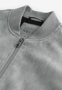 Veste en daim gris clair avec col côtelé et fermeture éclair argentée, taille moyenne, étiquette de marque "NEXT" visible à l'intérieur du col.
