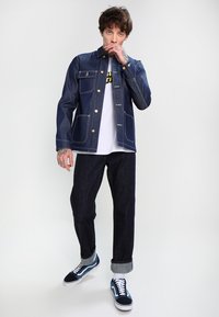 Dark blue denim jacket with contrasting yellow stitching, button closure, and two front pockets; paired with dark jeans and navy sneakers.