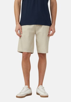 Mann trägt beige knielange Shorts, dunkelblaues T-Shirt und weiße Turnschuhe, steht vor einfachem Hintergrund.