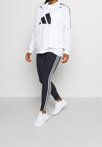 White zip-up jacket with black accents, white T-shirt featuring a large black logo, navy leggings with white stripes, and white athletic shoes.