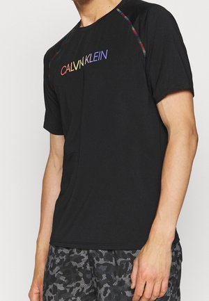 Homme portant un t-shirt noir à manches courtes Calvin Klein avec logo arc-en-ciel et surpiqûres, associé à un short camouflage noir et gris.