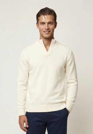 Hombre con cabello oscuro, vistiendo un suéter de punto color crema con cuello alto y pantalones azul oscuro, de pie contra un fondo claro.