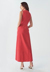 Červené maxi šaty bez rukávov s priliehajúcou sukňou, bočnými vreckami a hladkým povrchom. Modelka stojí v bielych sandáloch na vysokom opätku.