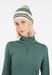 Beanie a righe verdi e bianche in maglia con una texture a coste e una piccola etichetta in pelle. Indossato con un top verde a collo alto con cerniera.