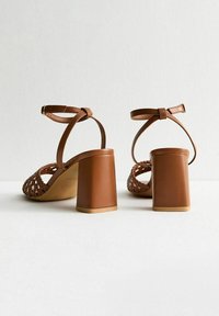 Brown high-heeled sandals with a woven upper design, adjustable ankle straps, and thick block heels. Smooth texture and matte finish.