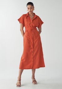 Red linen shirt dress with short sleeves, a belted waist, and front buttons. Features two patch pockets on the chest. Smooth texture.