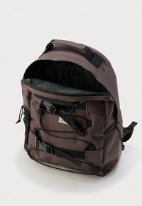 Καφέ σακίδιο με μαύρους ιμάντες και αγκράφες, μπροστινή τσέπη με φερμουάρ ανοιχτή για να δείχνει ευρύχωρο εσωτερικό με μαύρη επένδυση και δίχτυ στην πλαϊνή τσέπη.