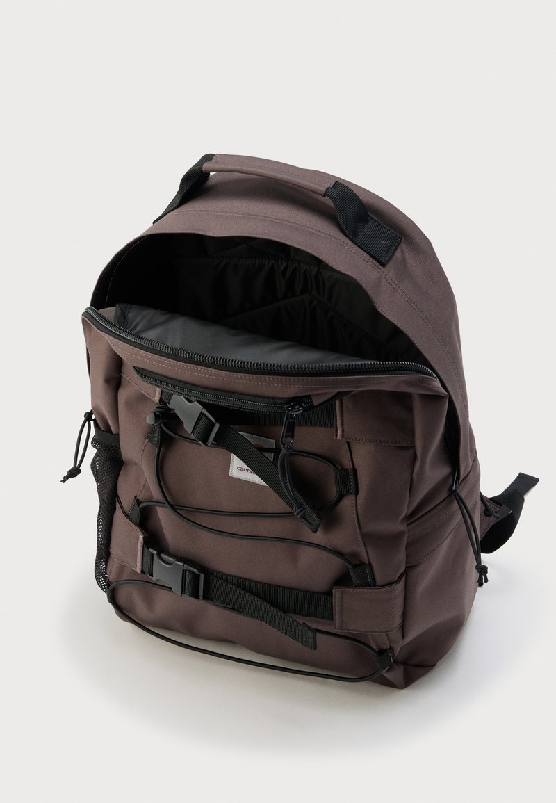 Bruine rugzak met zwarte riemen en gespen, ritsvak aan de voorkant open om de ruime, zwart gevoerde binnenkant en het zijvak van gaas te tonen.