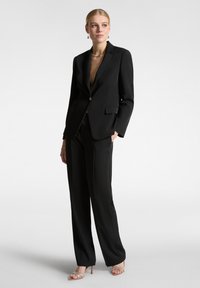 Blazer nero con rever eleganti, singolo bottone dorato e due tasche frontali, abbinato a pantaloni neri coordinati e sandali con tacco a stiletto.