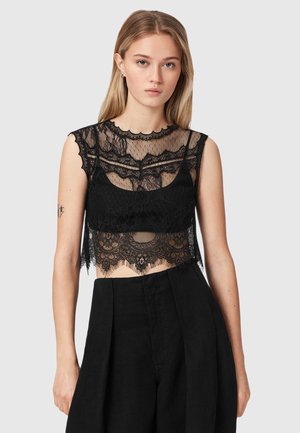 Jonge vrouw met lang blond haar draagt een zwarte kanten mouwloze cropped top en zwarte broek met hoge taille, staand tegen een effen achtergrond.