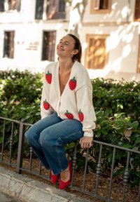Crème kleurige cardigan met aardbei-applicaties, gedragen over een diep uitgesneden top, gecombineerd met blauwe jeans en rode flats. Zittend op een hek te midden van het groen.