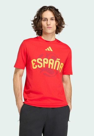 Jeune homme aux cheveux bouclés portant un t-shirt rouge avec le texte jaune "España" et le logo Adidas, debout les mains dans les poches de son pantalon noir.