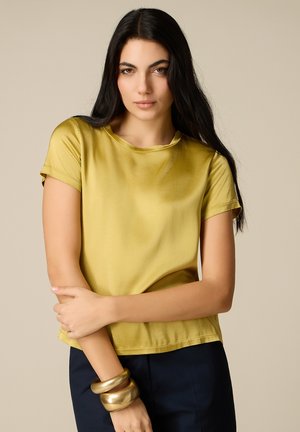 Kobieta z długimi czarnymi włosami, ubrana w żółtą satynową bluzkę z krótkim rękawem i ciemne spodnie, stojąca z założonymi ramionami i złotymi bransoletkami na nadgarstku.