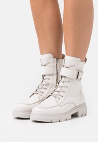 Bílé kotníkové kožené boty s lesklým povrchem, vybavené tkaničkami, bočním zipem a nastavitelným přezkovým páskem. Masivní gumová podrážka pro lepší trakci.