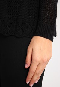 Musta neulepaita, jossa on teksturoitu kuosi, avoimia ompeleita ja aaltoileva helmataite. Käsivarsi lepää mustalla kangaspinnalla.