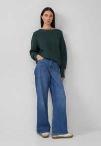 Donna che indossa un maglione a coste verde scuro, jeans a gamba larga blu e sneaker beige, in piedi davanti a una parete grigia chiara e uniforme.