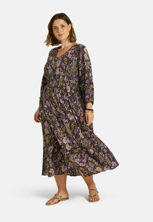 Vrouw draagt een lange, kleurrijke jurk met bloem- en paisleypatroon, lange mouwen en gouden sandalen, staand tegen een effen achtergrond.