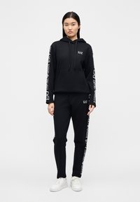 Γυναίκα που στέκεται φορώντας μαύρο φούτερ EA7 Emporio Armani με κουκούλα και ταιριαστό παντελόνι φόρμας με λευκά λογότυπα στα μανίκια και στα πόδια του παντελονιού, λευκά αθλητικά παπούτσια.