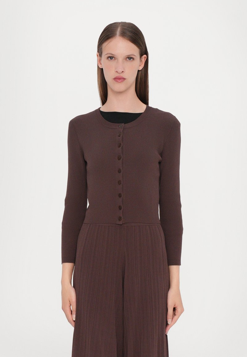 Cardigan marron à côtes avec de longues manches, col rond et fermeture à boutons sur le devant. Texture lisse, design ajusté, porté sur un haut noir.
