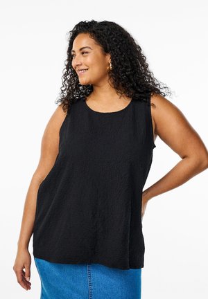 Smilende kvinde med krøllet hår iført en ærmeløs sort tekstureret top og blå denim nederdel, poserende med den ene hånd på hoften.