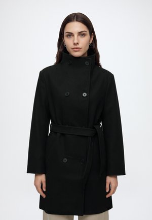Femme portant un manteau noir croisé avec un col montant et une ceinture, debout devant un fond blanc uni.