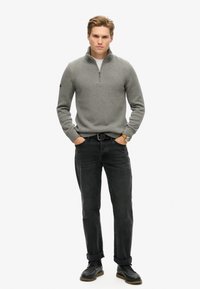 Grauer gestrickter Halbzip-Pullover mit geripptem Kragen, kombiniert mit schwarzen Jeans und schwarzen Schnürschuhen. Einfaches Design, keine sichtbaren Muster oder Logos.