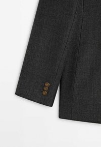 Manche de blazer gris foncé sur mesure avec trois boutons décoratifs en bronze sur le poignet, présenté sur fond blanc.