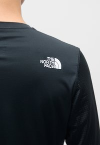 Svart atletisk skjorte med teksturert stoff, med et hvitt logo av The North Face på øvre del av ryggen, som fremhever det stilige designet.
