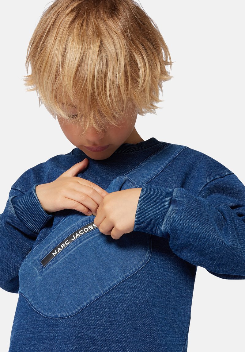 Sweatshirt bleu avec une grande poche avant ; présente une texture en denim et une étiquette noire proéminente portant la mention "MARC JACOBS." Coudes côtelés.