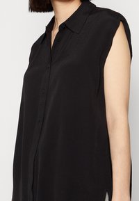 Ärmlös svart skjortblus med krage, bärs av en person med kort mörkt hår.