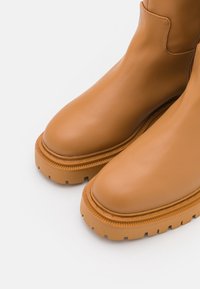 Botas de tobillo de cuero marrón claro con punteras redondeadas, suelas de goma texturizadas y detalles de costura en contraste a lo largo del borde de la suela.