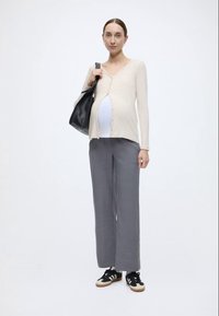 Μπεζ ζακέτα με κουμπιά, λευκό τοπ, γκρι παντελόνι με φαρδιά μπατζάκια και μαύρα αθλητικά παπούτσια με μπεζ λεπτομέρειες. Το μοντέλο κρατά μια μαύρη τσάντα tote.