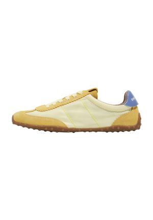Zapatilla deportiva de perfil bajo en amarillo y beige, con cordones blancos, suela de goma marrón con agarres redondos y una lengüeta azul en el talón con marca.