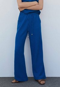 Personne portant un pantalon bleu ample avec un cordon de serrage à la taille et des sandales marron, se tenant les bras croisés contre un mur clair et uni.