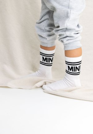 Chaussettes en coton blanches avec le texte "MINI" en noir et des accents rayés, portées avec un pantalon de survêtement gris, reposant sur un tissu texturé léger.