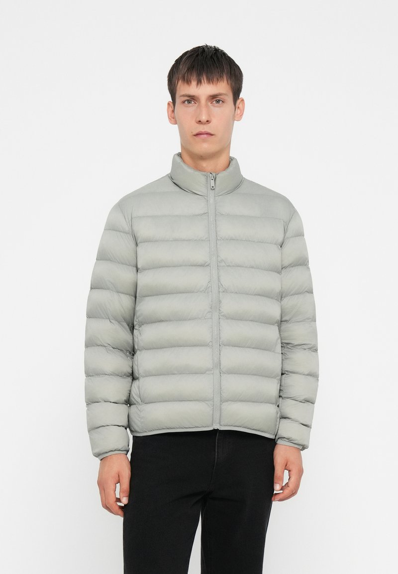 Lys grå pufferjakke med stående krage, glidelås foran og horisontal quiltet design. Stoffet virker glatt og polstret.