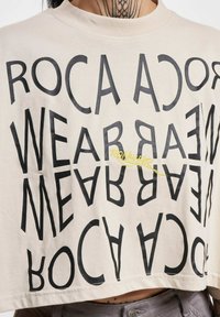 Beige cropped t-shirt featuring large, contrasting black text with an inverted design, highlighted with yellow stitching. Soft fabric texture.