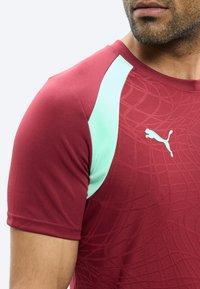 Mees, kes kannab burgundia värvi Puma spordisärki, millel on helesinised õlaaktsendid ja riidel peen muster, lõige nina tasemest allapoole.