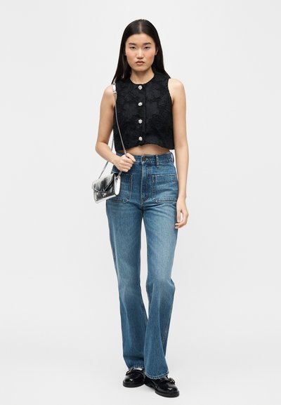 Νεαρή γυναίκα που στέκεται, φοράει μαύρο αμάνικο crop top με λουλουδάτο δαντελένιο σχέδιο, ψηλόμεσο μπλε τζιν, μαύρα μοκασίνια και ασημένια τσάντα ώμου.