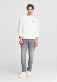 Maglione bianco con colletto rotondo, testo del logo nero sul petto, abbinato a jeans grigi chiari e sneakers bianche. Texture liscia, vestibilità casual.