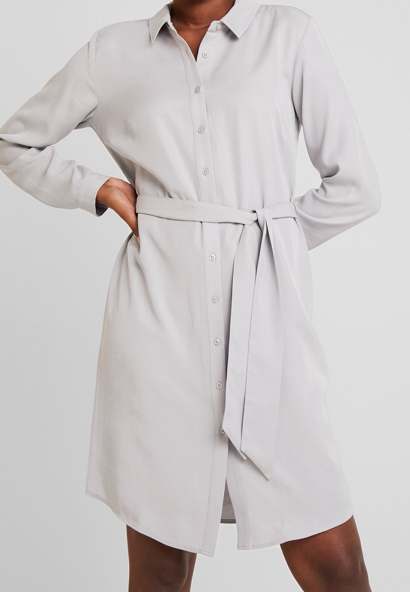 Vestito camicia grigio chiaro a maniche lunghe con apertura con bottoni, colletto e cintura in tessuto che stringe in vita. Texture liscia, lunghezza al ginocchio.